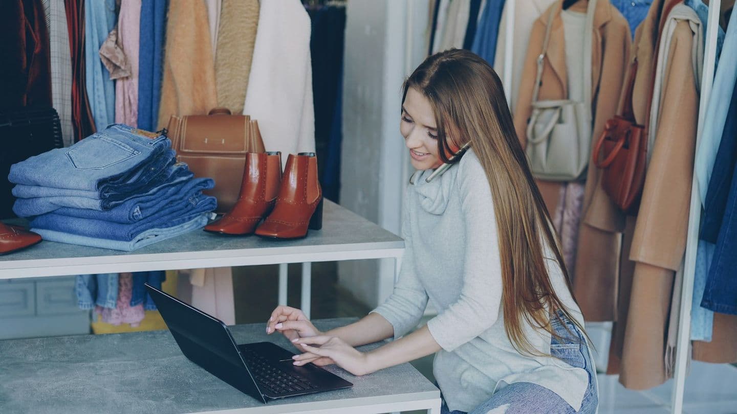 Una joven empresaria trabaja con un portátil en su tienda de ropa, rodeada de prendas, bolsos y calzado.